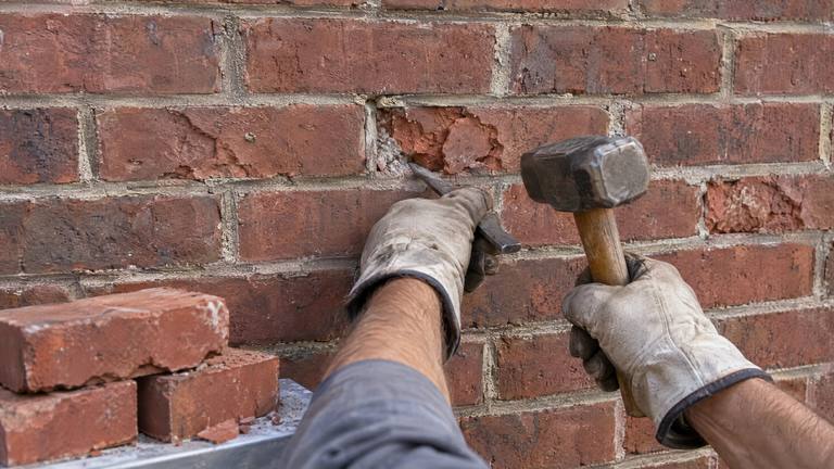 Gloved hands work a cold chisel and a lump hammer into the mortar around spalled red bricks with flaking faces.