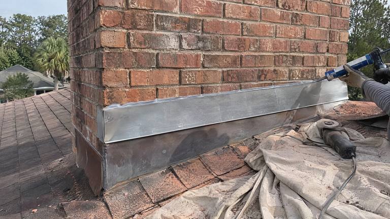 A bead of sealant goes along the top edge of new metal flashing at the base of a red brick chimney.