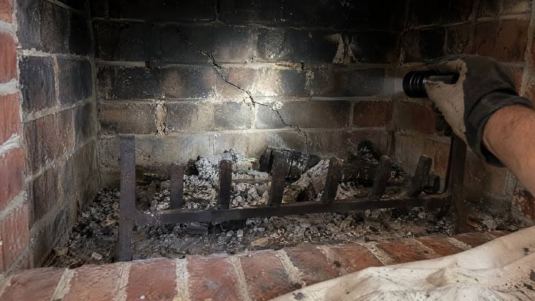 A gloved hand shines a flashlight on a long crack in the sooty back wall of a brick firebox, behind a cast iron grate and ash.