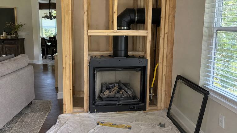 A direct-vent gas fireplace unit sits in a framed wall with its black vent pipe running up through the framing.