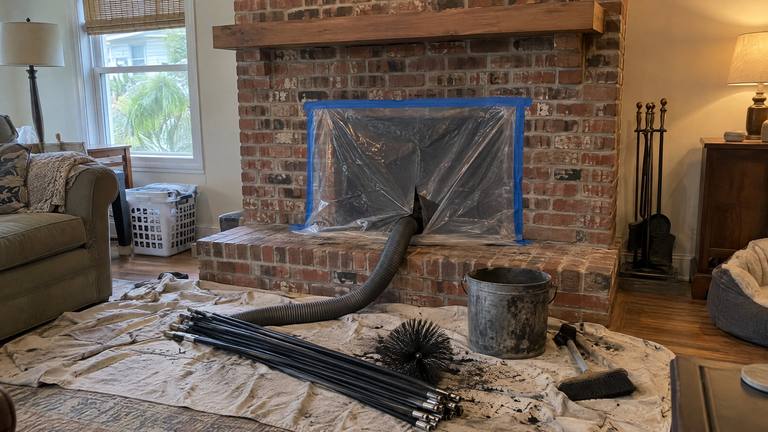 A brick fireplace sealed with plastic sheeting and a vacuum hose, with sweep rods, a sooty chimney brush and an ash bucket on a drop cloth.