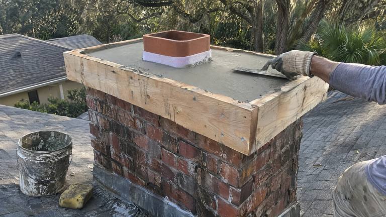 Wood form boards frame the top of a red brick chimney while wet concrete is troweled into a new crown.