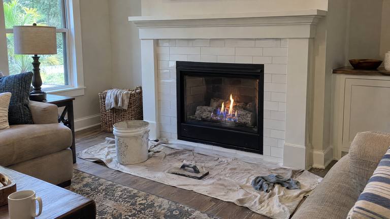 A newly installed gas fireplace burns behind glass in a fresh white tile surround, with a bucket and tools on a drop cloth.