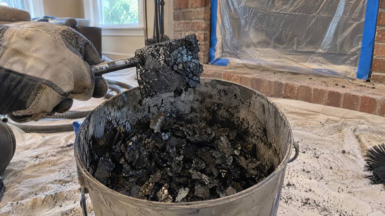 Shiny black flakes of glazed creosote scraped from a flue lie in a metal bucket beside a scraping tool.