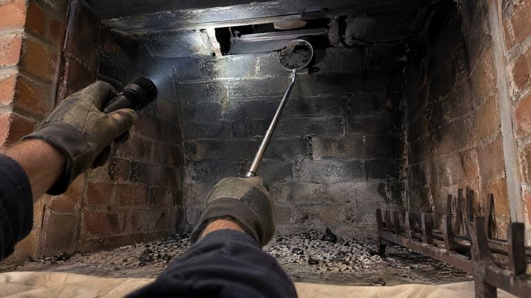 Gloved hands hold a flashlight and a small inspection mirror inside a sooty brick firebox, angled up toward the flue.