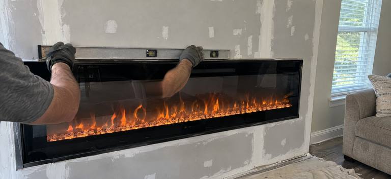 Gloved hands set a level on top of a long linear electric fireplace built into a new wall of unfinished drywall.