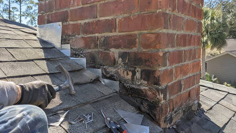 New aluminum step flashing pieces are woven under each shingle course along the side of a red brick chimney.