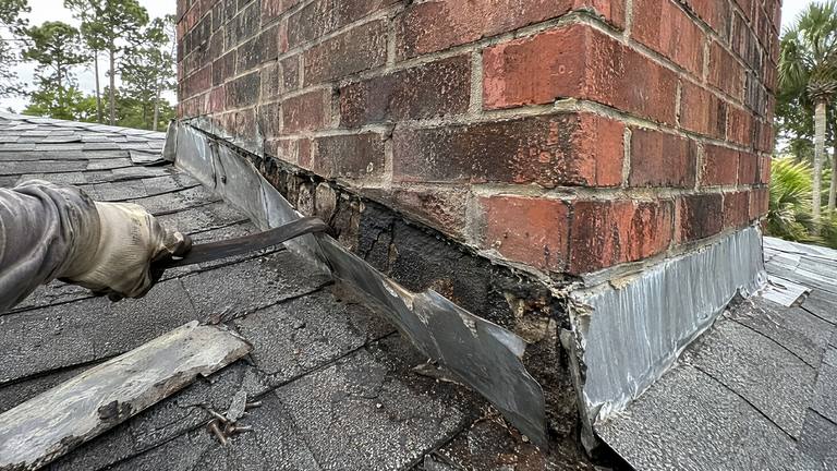 A gloved hand pries old metal flashing away from the base of a brick chimney, exposing dried roofing tar above the shingles.