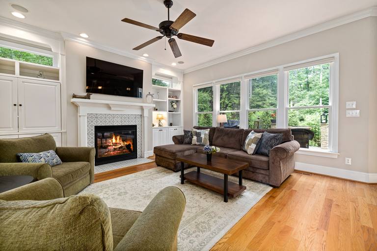 A family room with a lit fireplace in a patterned tile surround under a white mantel, a ceiling fan, a brown sectional sofa and green armchairs.