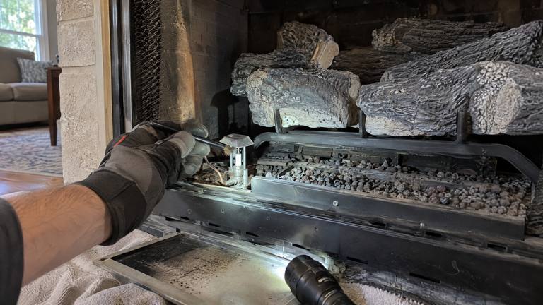 A gloved hand cleans the pilot assembly of a gas fireplace with its glass front off and logs moved aside.