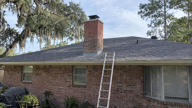 A red brick chimney with a plain black cap rises from the gray shingle roof of a brick ranch house, a ladder at the eave.