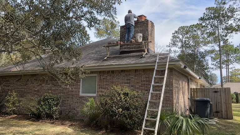 A worker on a roof resets loose bricks at the top of a red brick chimney with a trowel and a bucket of mortar.