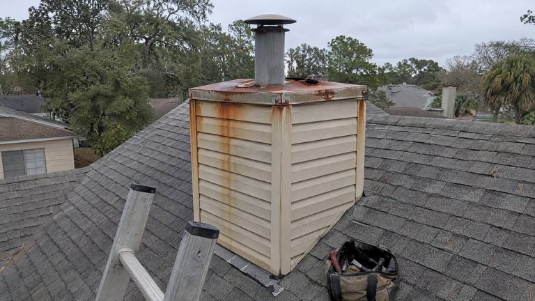 A rusted chase cover with a round flue pipe tops a sided chimney chase, with rust streaks down the siding and a ladder at the roof edge.