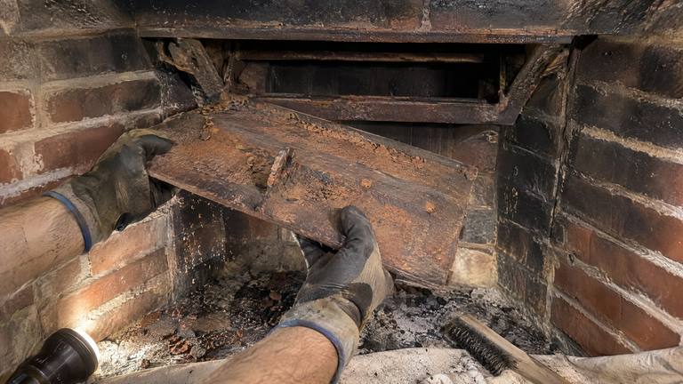 Gloved hands lift a rusted cast iron damper plate out of the throat of a sooty brick fireplace.