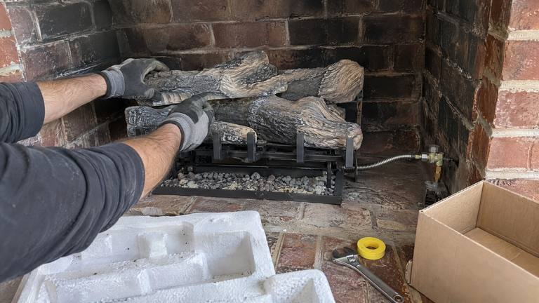 Gloved hands set ceramic gas logs on a grate over the burner in a soot-darkened brick firebox, with a wrench and thread tape on the hearth.
