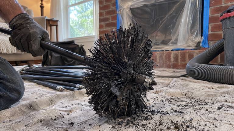 A round wire chimney brush caked with black soot rests on sweep rods on a drop cloth in front of a fireplace.