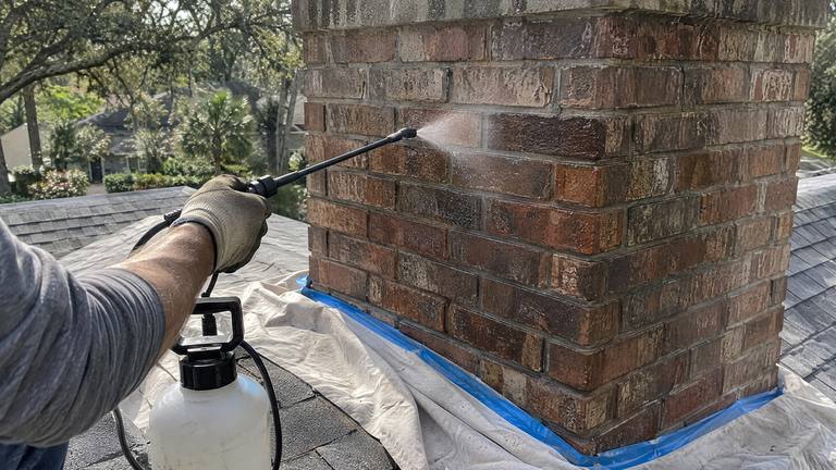 The wand of a pump sprayer wets the side of a red brick chimney with clear sealer, the treated band darker.