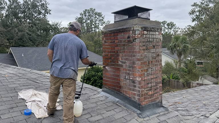 A worker on a roof sprays clear sealer from a pump sprayer onto the side of a weathered red brick chimney.