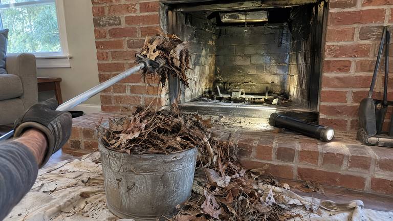 A bucket of leaves and twigs pulled from a chimney sits on a drop cloth in front of a brick fireplace.