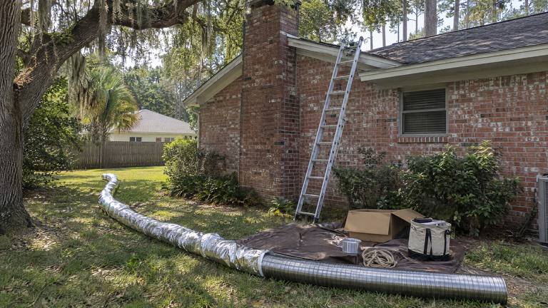 A long flexible stainless steel chimney liner lies on the lawn beside a ladder set against a brick house.