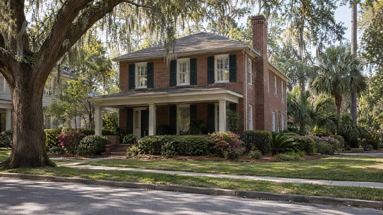 A two-story red brick house with a front porch and a tall side chimney on a street lined with large live oaks.