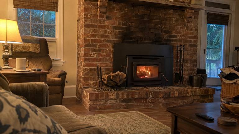 A small fire burns behind the glass of a black wood-burning insert set in a soot-stained red brick fireplace, with a log rack on the hearth.
